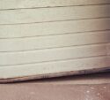 A garage door is dented and bent at the bottom, with visible dirt on the ground in front of it.