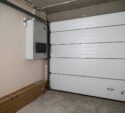 Closed white sectional garage door in a bare concrete garage with an electrical panel and some long, boxed items on the floor.