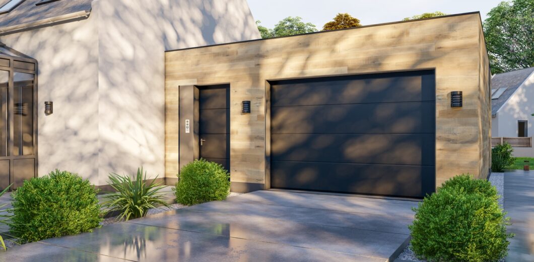 Modern detached garage with light wood siding, a dark single garage door, side entry door, two wall lights, and landscaped bushes on a concrete driveway.