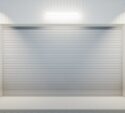 Closed white industrial garage door with protective yellow-and-black striped bollards on either side, illuminated by a wall-mounted light and surrounded by large windows.