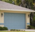 A closed light blue garage door on a beige house with two outdoor wall lights and greenery surrounding the driveway.