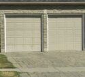 Two closed beige garage doors on a stone-faced building with a paved driveway and some patches of grass in the foreground.