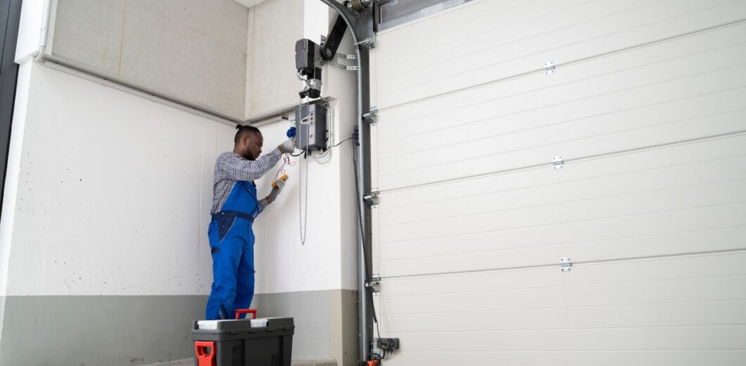 A technician in blue overalls repairs or maintains an electric garage door opener, standing next to a large closed garage door with a toolbox on the ground.