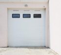 Closed white garage door with three rectangular windows near the top, set in a light-colored exterior wall on a concrete driveway.