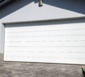 A closed white sectional garage door on a modern house with a paved driveway and a small evergreen shrub in the foreground.
