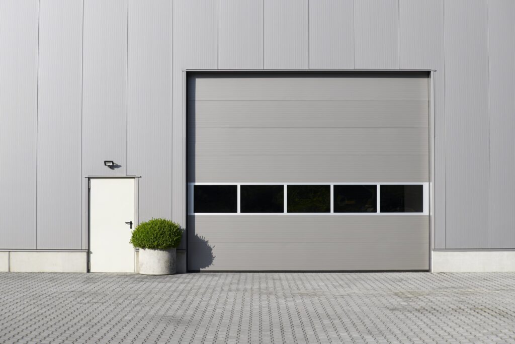Gray industrial building exterior with a large closed garage door, a small side door, a wall-mounted light, and a potted shrub on a paved surface.