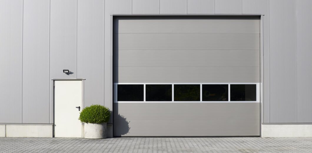 Gray industrial building exterior with a large closed garage door, a small side door, a wall-mounted light, and a potted shrub on a paved surface.
