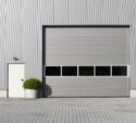 Gray industrial building exterior with a large closed garage door, a small side door, a wall-mounted light, and a potted shrub on a paved surface.