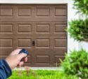 A person holds a remote control in front of a closed brown garage door, with greenery visible in the foreground.