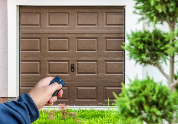 A person holds a remote control in front of a closed brown garage door, with greenery visible in the foreground.