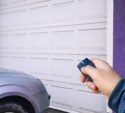 A person uses a remote control to open a closed white garage door next to a parked silver car.