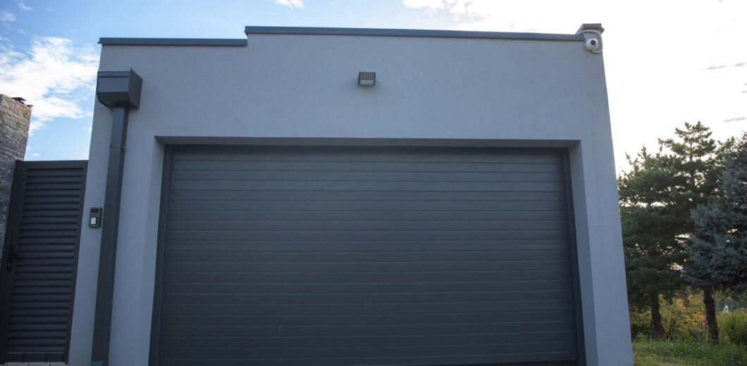 A modern gray garage with a closed overhead door, an outdoor light above, and trees visible to the right under a partly cloudy sky.