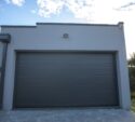 A modern gray garage with a closed overhead door, an outdoor light above, and trees visible to the right under a partly cloudy sky.
