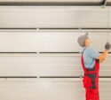 A worker in red overalls and a cap is pulling a cord to open or close a large white garage door.