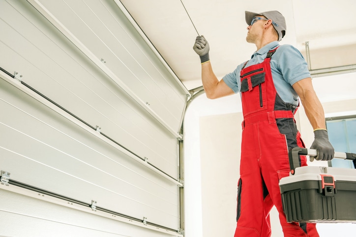 A technician in red overalls and gloves repairs a garage door while holding a toolbox.