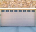 Closed beige garage door set in a stone wall facade with small windows at the top and two black lantern-style wall lights on either side.