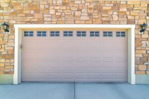 Closed beige garage door set in a stone wall facade with small windows at the top and two black lantern-style wall lights on either side.