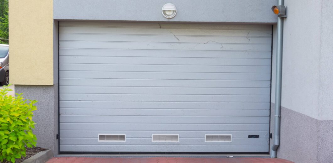 Gray metal garage door with ventilation panels, set in a light gray and yellow building, next to a green shrub and red brick pavement.