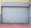 Gray metal garage door with ventilation panels, set in a light gray and yellow building, next to a green shrub and red brick pavement.