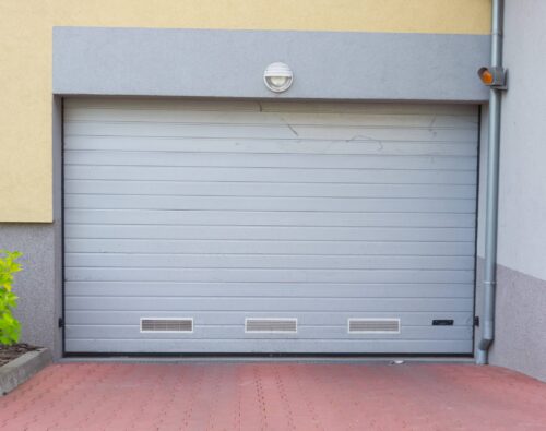 Gray metal garage door with ventilation panels, set in a light gray and yellow building, next to a green shrub and red brick pavement.