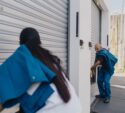 Two workers in blue uniforms are opening or closing metal roll-up doors on a loading dock or storage facility.