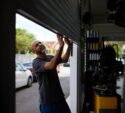 A man stands inside a garage, manually lifting a metal roll-up door. Cars and trees are visible outside in the background.