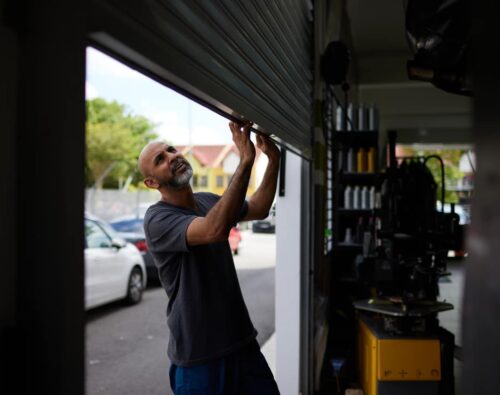 A man lifts a metal garage door inside an automotive workshop, with parked cars and buildings visible outside in daylight.