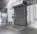 Two gray industrial roll-up doors in a warehouse setting, with wall-mounted control panels, a fire extinguisher, and utility fixtures nearby.