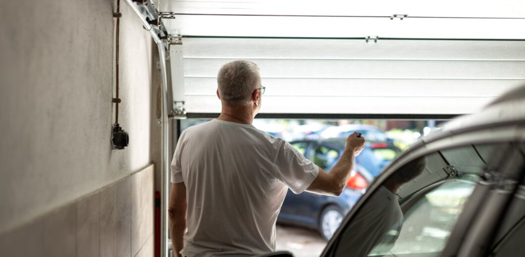 A man in a white t-shirt stands in a garage, holding a remote and opening the garage door, with parked cars visible outside.