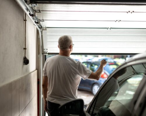 A man in a white t-shirt stands in a garage, holding a remote and opening the garage door, with parked cars visible outside.