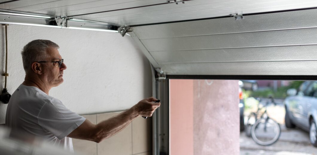 A man in a white t-shirt uses a remote control to open a garage door, with a bicycle and car visible outside.