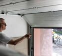 A man in a white t-shirt uses a remote control to open a garage door, with a bicycle and car visible outside.