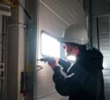 A worker wearing a hard hat uses a power drill to install or repair a metal door inside an industrial building.