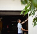 A man in a light blue shirt stands outside a garage, reaching up to close or open the garage door. A black vehicle is parked inside. Green leaves are visible in the foreground.