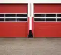 Two closed red garage doors with horizontal black and white stripes, set in a white and red building, viewed from a concrete driveway.