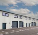 A row of modern industrial units with large roller doors and parking spaces marked in front, under a partly cloudy sky.