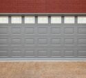 A closed gray garage door with small rectangular windows at the top, set in a red brick wall with two outdoor wall lights on either side.