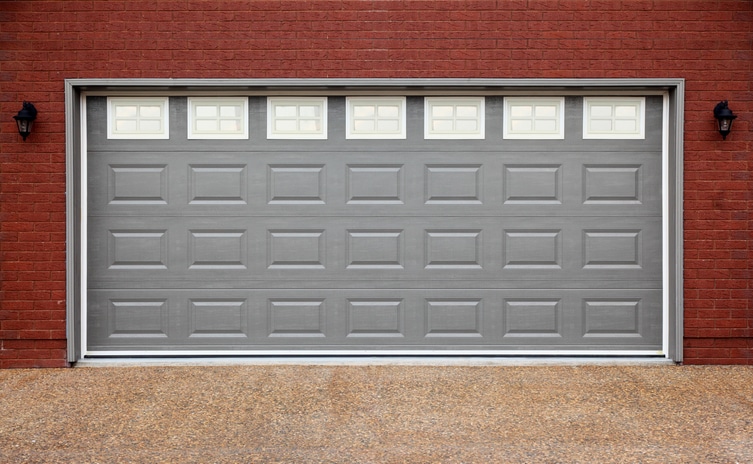 A closed gray garage door with small rectangular windows at the top, set in a red brick wall with two outdoor wall lights on either side.