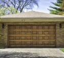 A brick house with a closed wooden garage door, two outdoor wall lanterns, and a paved driveway surrounded by trees and shrubs.