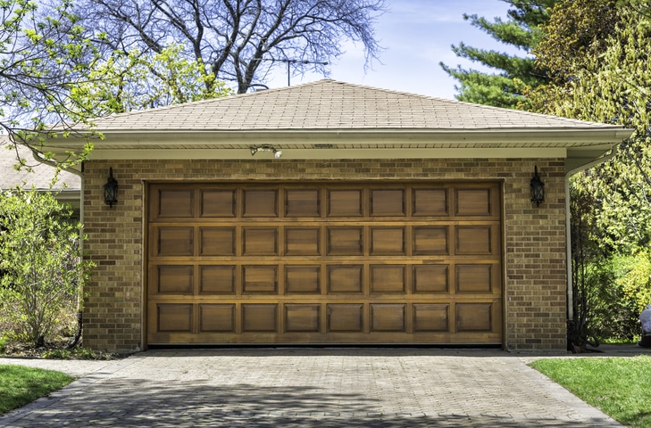 A brick house with a closed wooden garage door, two outdoor wall lanterns, and a paved driveway surrounded by trees and shrubs.