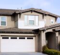 Two-story suburban house with beige stucco exterior, white trim, a double garage door, and landscaped front yard.