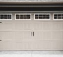 Closed beige garage door with rectangular panel design and four windows at the top, featuring black decorative handles and hardware.