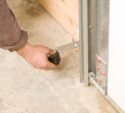 A person’s hand adjusts a metal component on a garage door track near the concrete floor.