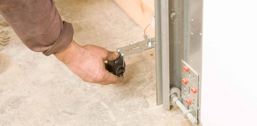 A person adjusts the sensor near the bottom of a garage door track, with visible wiring and mounting hardware on a concrete floor.