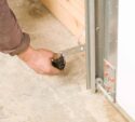 A person adjusts the sensor near the bottom of a garage door track, with visible wiring and mounting hardware on a concrete floor.