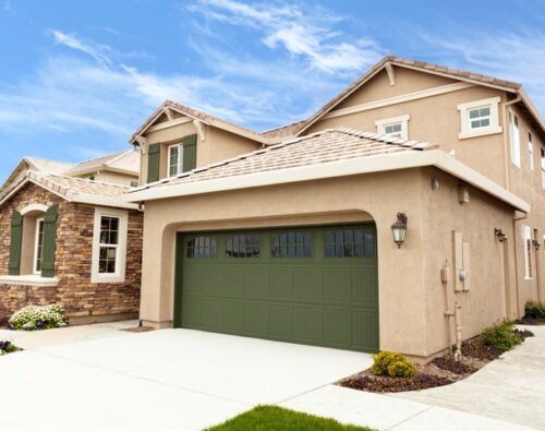 Two-story beige house with a green garage door, stone accents, and a manicured front yard under a blue sky.
