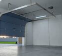A modern, empty garage with a partially open blue sectional garage door, concrete floor, and white walls.