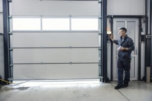A man in work clothes stands by a control panel next to a large closed garage door in an industrial setting.