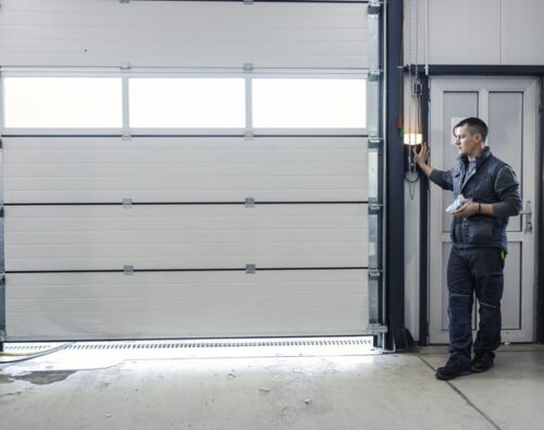 A man in work clothes stands by a control panel next to a large closed garage door in an industrial setting.