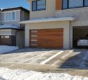 A modern two-story house with a wooden garage door and a car parked inside the garage. The driveway and sidewalk are partially cleared of snow.